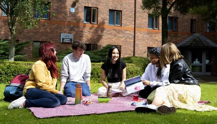 A group of students sit outside their halls on a blanket, enjoying a picnic together.
