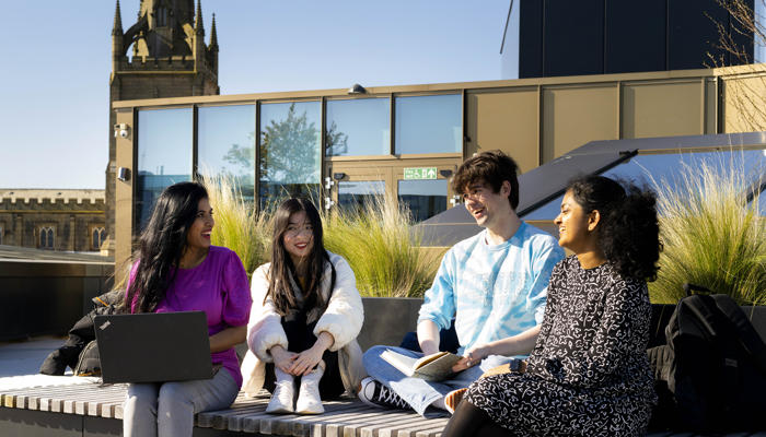 four international students sat on the roof terrace of the student centre on a bench on a sunny day